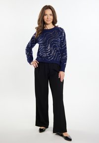 Marineblauer Pullover mit metallischen Wellmustern, strukturiertem Stoff und gerippten Bündchen, kombiniert mit weit geschnittenen schwarzen Hosen und glänzenden schwarzen Schuhen.