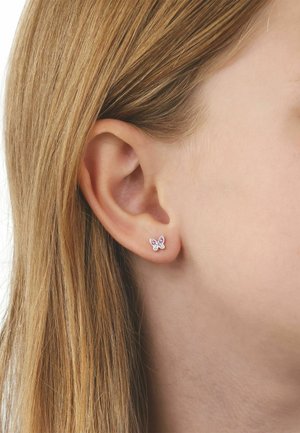 Primer plano de una oreja izquierda con cabello rubio liso, que lleva un pequeño pendiente de botón en forma de mariposa con piedras preciosas rosas y transparentes.