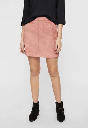 Femme portant un haut noir à manches longues, une mini-jupe en daim rose et des bottines noires avec des brides cloutées, debout devant un fond uni.