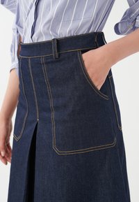 Denim krilo v temno modri barvi, z dvema velikima sprednjima žepoma z rumenim šivanjem in sprednjim razporkom.