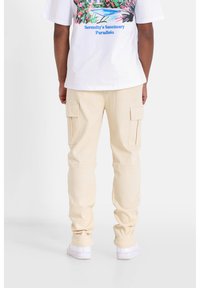 Beige cargobroek met een relaxte pasvorm, twee zijzakken en zichtbare stiksels, gecombineerd met een wit graphic t-shirt. Witte sneakers.