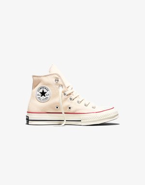 Beige színű magas szárú Converse Chuck Taylor All Star tornacipő fehér fűzőkkel, fekete csillag logóval, valamint piros és fekete talpsávokkal.