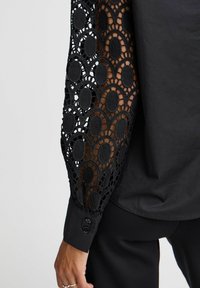 Schwarze Bluse mit aufwendigen langen Spitzenärmeln mit ovalen Mustern, solidem schwarzen Stoff im Torso und einem geknöpften Manschetten-Detail.