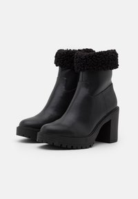 Schwarze Lederstiefeletten mit strukturiertem schwarzem Kunstfellbund, breitem hohen Absatz und geriffelter Sohle für besseren Halt. Glatte Oberfläche mit minimalen Nähten.