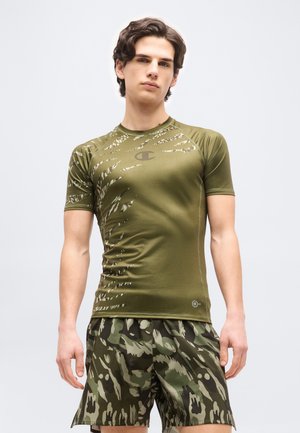 Mladý muž oblečený v priliehavom olivovo zelenom športovom tričku s pruhmi podobnými tigrej srsti a maskáčových kraťasoch, stojaci pred jednoduchým pozadím.