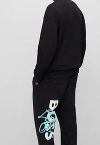 Conjunto de sudadera y pantalones deportivos negros. La sudadera tiene cuello redondo; los pantalones presentan un diseño gráfico azul y blanco en el muslo. Tela de algodón suave.