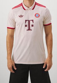 Beżowa koszulka polo z czerwonymi akcentami, z logo FC Bayern Monachium, marką Adidas i teksturowym wzorem na materiale.