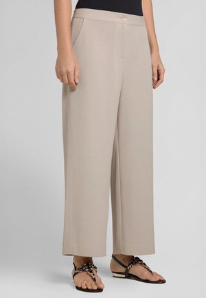 Pantaloni beige a gamba larga con tasche laterali, vita alta e chiusura con bottoni, indossati con sandali bassi neri decorati con fiori.