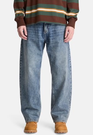 Person iført løstsiddende, falmede blå jeans, brun og grønstribet langærmet skjorte og tan-farvede arbejdstøvler, står mod en ensfarvet baggrund.