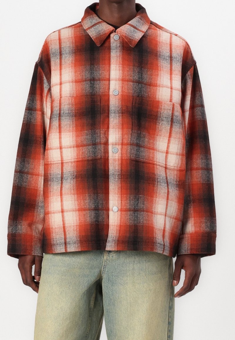 Homme portant une chemise à carreaux rouge, noire et blanche à boutons avec col et un jean bleu clair sur fond blanc.