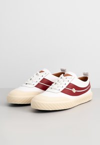 Paire de baskets blanches avec semelles en caoutchouc beige et bandes rouges sur le côté, avec des lacets blancs et un petit logo doré « B » sur le côté.