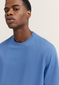 Blue cotton t-shirt with a round neck, short sleeves, and a smooth texture. Subtle branding near the hem. Simple, casual design.