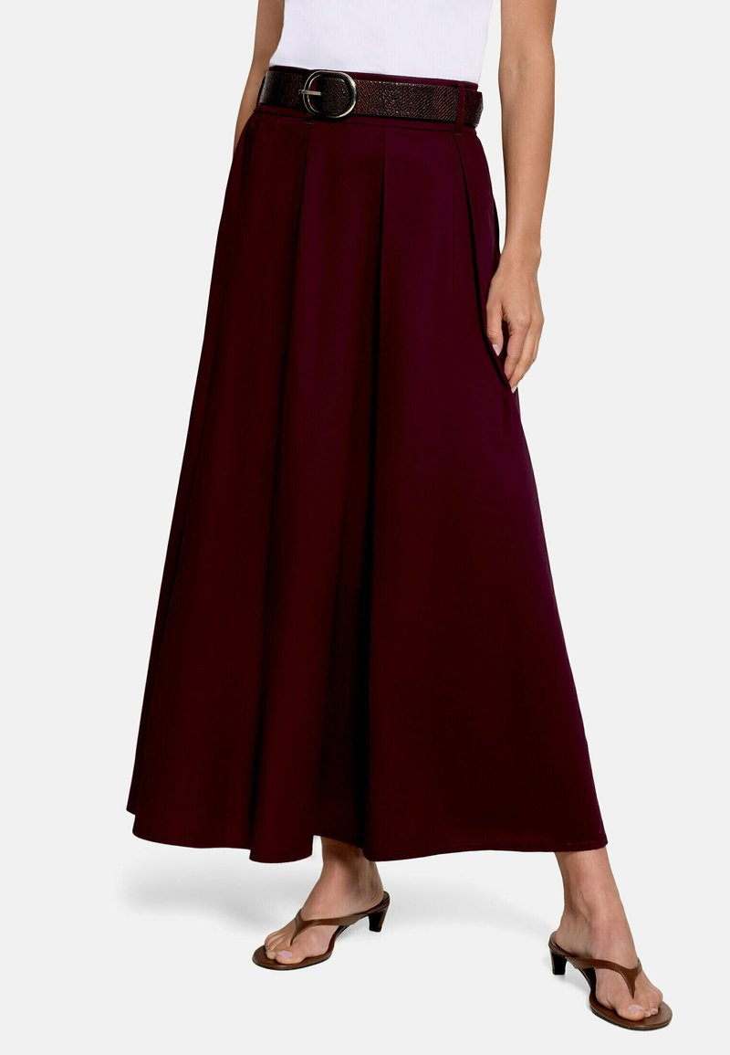 Femme portant une jupe large plissée de couleur bordeaux foncé, associée à un haut blanc, une ceinture noire et des sandales marron à talons.