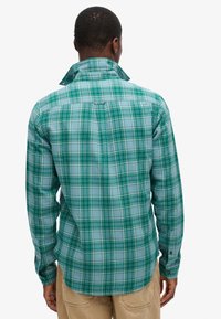 Camicia a quadri verde e blu con colletto abbottonato, maniche lunghe e orlo curvo. Presenta una cucitura a yoke sul retro. Materiale in misto cotone.