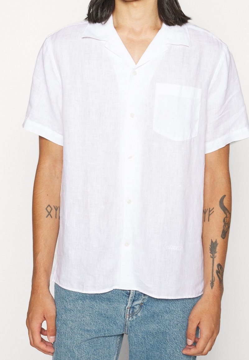 Personne portant une chemise blanche à manches courtes avec une poche poitrine et un jean en denim bleu clair, montrant des tatouages sur les deux avant-bras.
