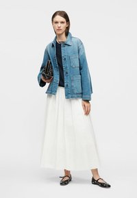 Γυναίκα που φορά τζιν μπουφάν, μαύρο τοπ, λευκή πλισέ φούστα, μαύρες μπαλαρίνες και κρατά ένα μαύρο τσαντάκι με καρφιά.