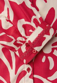 Poignet de vêtement en tissu à motif floral rouge et blanc, avec deux boutons rouges et un bord en dentelle sur une manche.