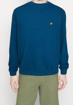 Homme portant un sweat-shirt à manches longues bleu marine uni avec un petit logo d'oiseau jaune sur la poitrine et un pantalon vert olive.
