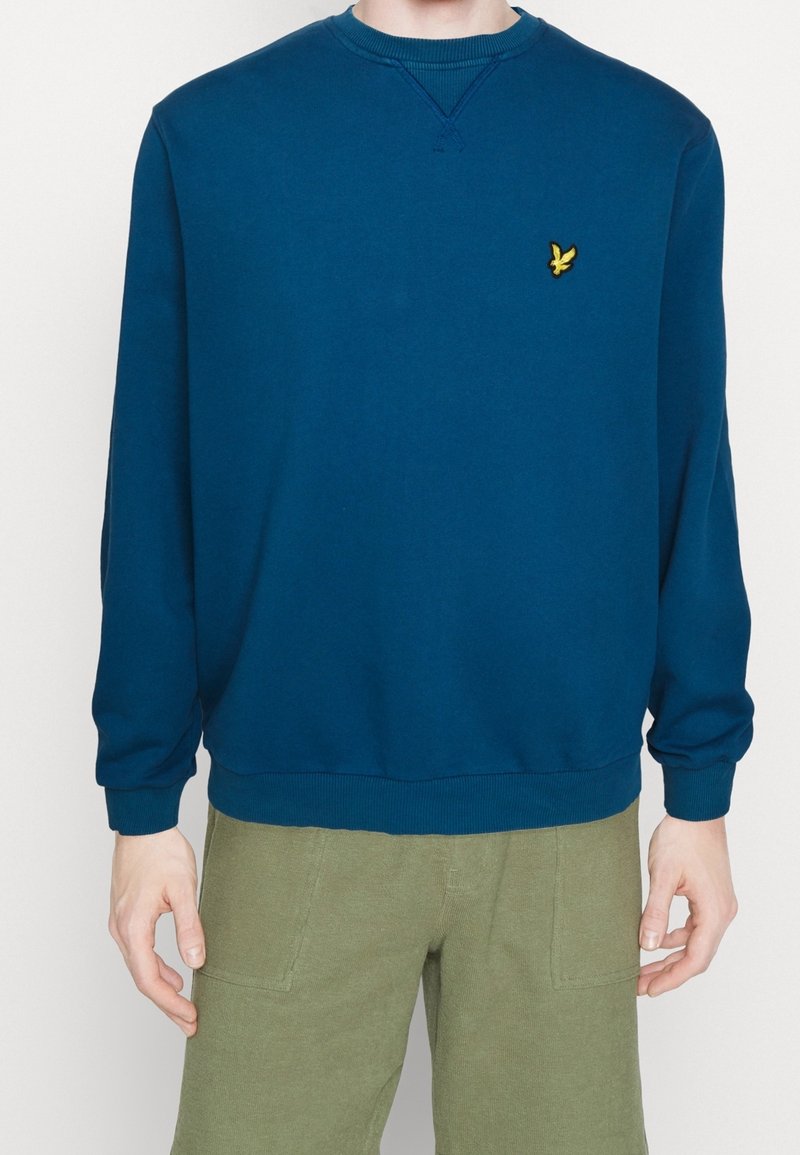 Homme portant un sweat-shirt à manches longues bleu marine uni avec un petit logo d'oiseau jaune sur la poitrine et un pantalon vert olive.