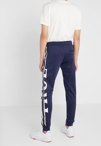 Mörkblå träningsbyxor med vita och beige sidostriper och text. Mjuk tyg, elastisk midja, smalnande ben och sidoficka.