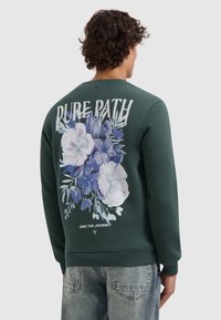 Groene sweatshirt met ronde hals, voorzien van een bloemenprint in blauw- en wittinten op de achterkant met de tekst "PURE PATH" erboven en "OWN THE JOURNEY" eronder.
