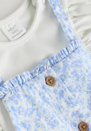 Biała bawełniana kombinezon z krótkimi falbankowymi rękawami, pod niebieskim dżinsowym ogrodnikiem w kwiatowy wzór z dwiema okrągłymi drewnianymi guzikami.