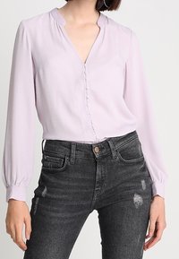 Blusa lavanda chiara con scollatura a V, chiusura frontale con bottoni e maniche lunghe a sbuffo, abbinata a jeans neri strappati.