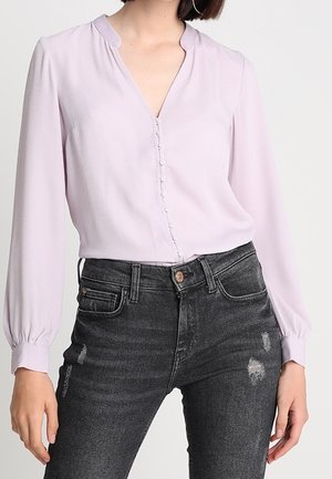 Blusa de lavanda clara con escote en V, cierre de botones en la parte frontal y mangas largas abullonadas, combinada con jeans negros desgastados.