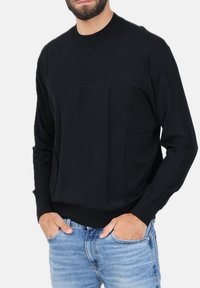 Maglione nero lavorato a maglia con scollo rotondo, caratterizzato da un pannello frontale testurizzato e polsini a costine, indossato con jeans in denim azzurro chiaro.