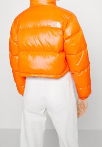 Krótkia puchowa kurtka w błyszczącym pomarańczowym kolorze z przeszywanymi panelami i wysokim kołnierzem. Z białym logo na lewym ramieniu. Noszona z białymi spodniami.