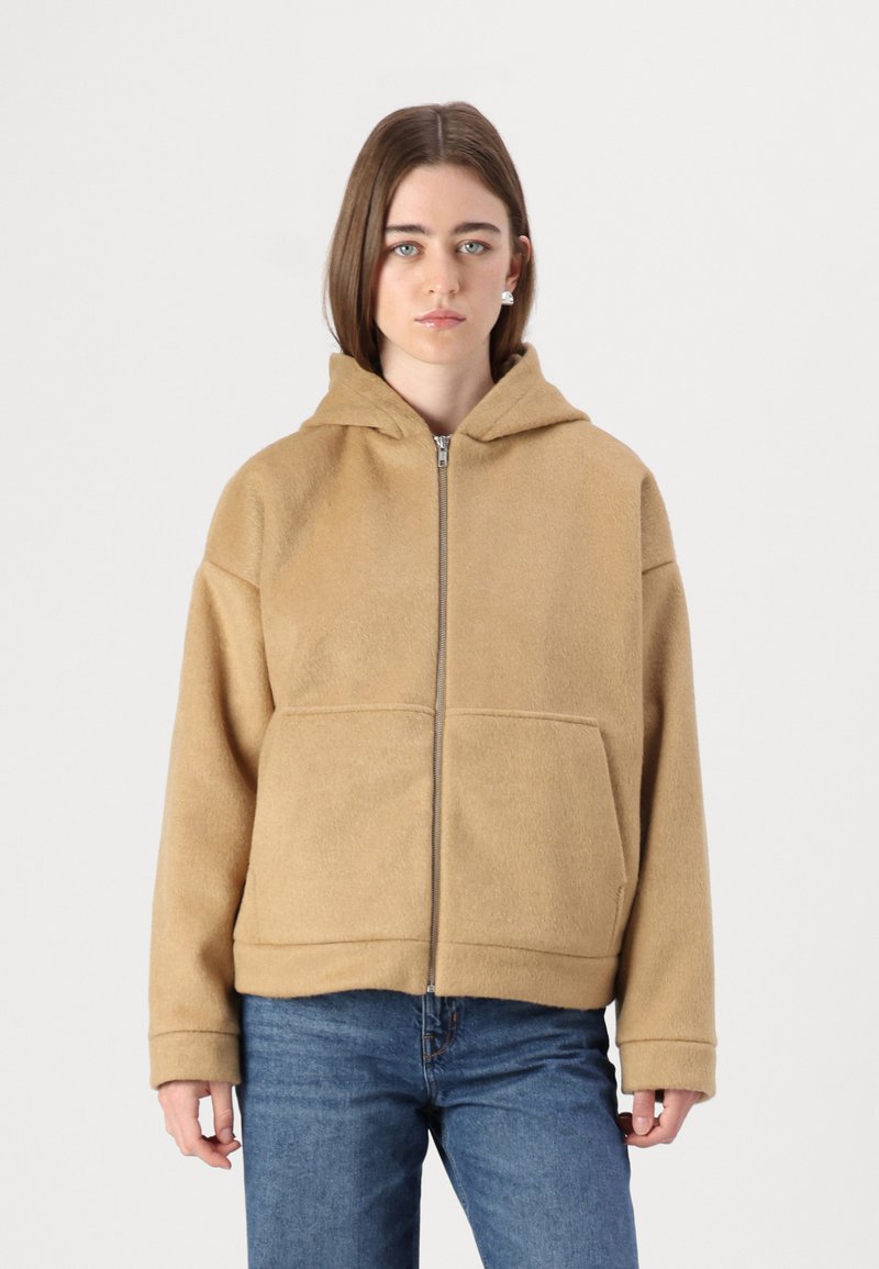Beige hettejakke i myk, teksturert stoff, med glidelås foran og to store lommer. Løs passform med ribbestrikkede mansjetter.