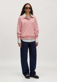 Lichtroze oversized sweatshirt met een kraag en knoopdetails, gelaagd over een wit met zwart gestreept shirt, gecombineerd met brede donkere jeans.