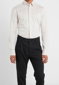 Camicia a maniche lunghe color beige chiaro con colletto abbottonato, abbinata a pantaloni neri con vita elastica e frontale plissettato.