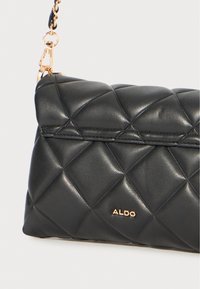 Borsa a tracolla in pelle trapuntata nera con catena. Presenta un accento con logo dorato e una forma strutturata. Texture liscia con una finitura lucida.
