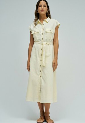 Mujer con vestido midi crema sin mangas con botones, bolsillos en el pecho y cinturón en la cintura, combinado con sandalias marrones con tiras.