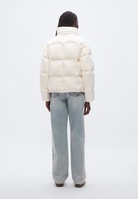 Κρεμ μπουφάν τύπου puffer με ψηλό γιακά και σχεδιασμό καπιτονέ, σε συνδυασμό με ανοιχτό μπλε τζιν παντελόνι φαρδύ, εμφανιζόμενα από πίσω.