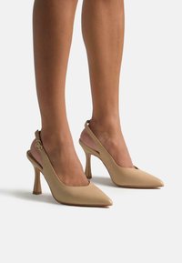 Tacones slingback en color nude con punta afilada y un tacón de aguja delgado de altura media. Cuero suave con un acento de ante suave en la parte trasera.