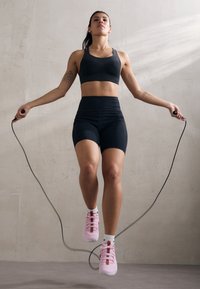 Sort sports-bh og højtaljede shorts med et struktureret design. Hopper med sjippetov, iført lyserøde sneakers og hvide sokker på et glat gulv.