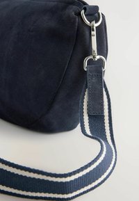 Esquina de bolso de ante azul marino con broche plateado que sujeta una correa de hombro de tela a rayas azules y blancas.