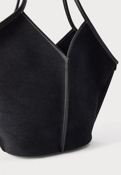 Sac à main noir texturé avec des côtés structurés et anguleux ainsi que des poignées arrondies, comportant des coutures claires visibles sur les bords, sur un fond uni.