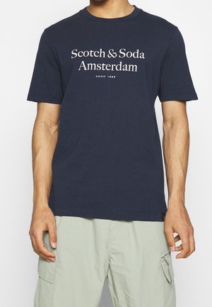 Homme portant un t-shirt bleu marine avec le texte « Scotch & Soda Amsterdam Anno 1985 » et un short cargo beige clair sur fond blanc.