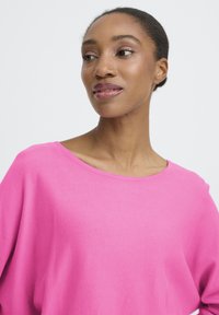 Femme à la peau foncée et aux cheveux attachés, portant un haut rose vif, regardant vers sa gauche devant un fond blanc uni.