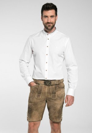 Weißes Langarmhemd mit hohem Kragen und braunen Lederhosen, die mit aufwendiger Stickerei und Knopfakzenten verziert sind. Schwarzer Gürtel mit einem Hirschmotiv.