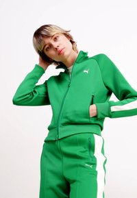 Green Puma tracksuit made of smooth material, featuring a zip-up jacket with white side stripes and matching pants, showcasing a modern fit.