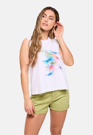 Mujer con cabello largo y ondulado que lleva una camiseta blanca sin mangas con un estampado colorido de libélula y pantalones cortos verde claro remangados.