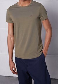 Oliivinvihreä lyhythihainen t-paita, valmistettu pehmeästä kankaasta, jossa pyöreä pääntie ja pieni logo vasemmassa rinnassa. Yhdistetty tummansinisiin housuihin.