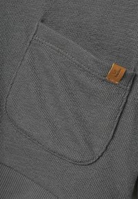 Tela texturizada gris con un bolsillo de parche y una etiqueta de cuero marrón. La superficie tiene un patrón sutil y en relieve para mayor detalle.