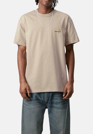 Camiseta de algodón beige con corte relajado, mangas cortas y un logotipo de Carhartt bordado en marrón en el lado izquierdo del pecho. Lleva puesta con jeans azules.