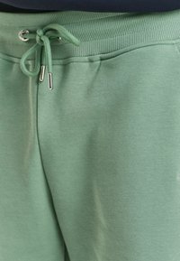 Pantalons de survêtement vert clair avec une taille à cordon de serrage, texture côtelée au niveau de la taille et finition lisse sur le tissu. Accents argentés sur le cordon.