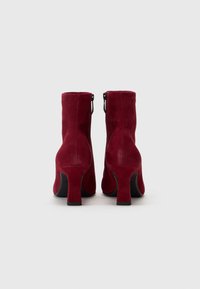 Stivaletti alla caviglia in suede rosso con una texture liscia, chiusura con zip e un tacco robusto. Il design presenta una punta arrotondata e dettagli di cuciture sottili.